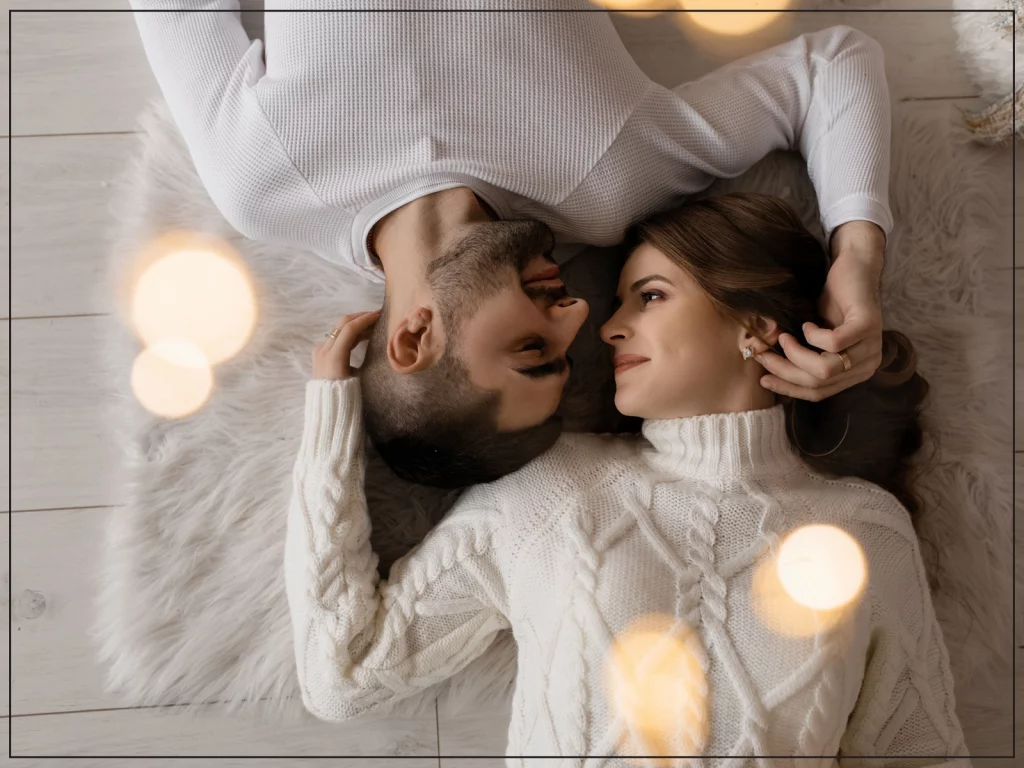 Man and woman wearing white cardigans laying on the floor looking into each other's eyes.