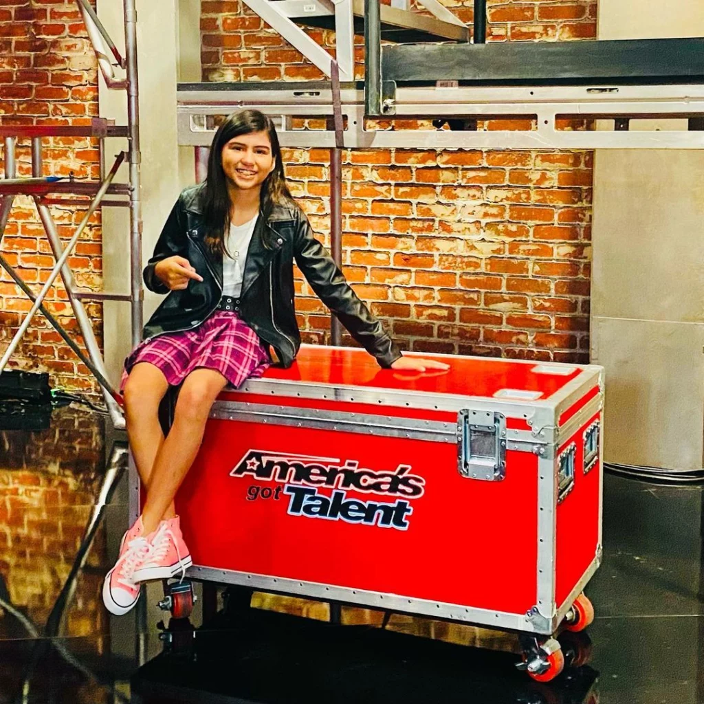 Madison wearing a red skirt and black jacket, sitting on the red trunk.